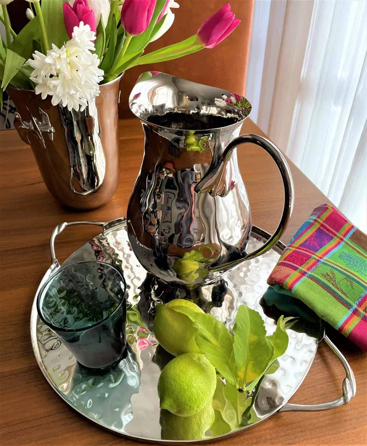 Polished silver pitcher, tray, and tumbler arranged with pink tulips, green fruit, and a plaid napkin